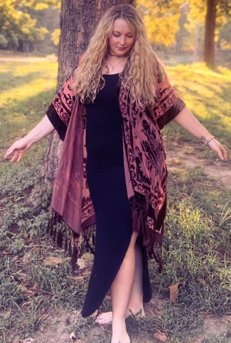 Rebekah standing grounded against a tree, arms raised, in a black dress and rose-patterned kimono