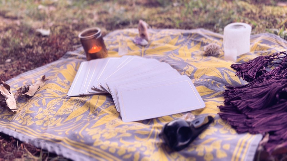 A fanned deck of plain gold-edged cards on a gold and purple blanket, with a candle, crystals and a raven-skull pipe