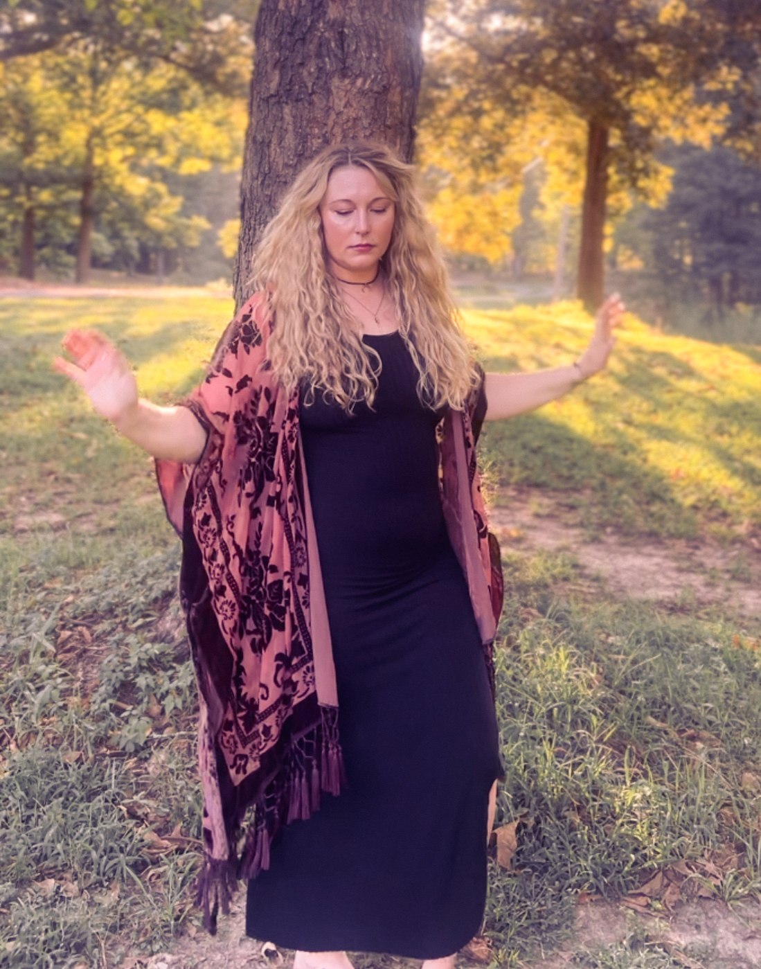 Rebekah standing grounded against a tree, arms raised, in a black dress and rose-patterned kimono